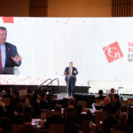 A speaker stands on stage at the Sports Betting Executive Summit, with a large screen displaying the speaker's image behind them. The audience is seated in rows, facing the stage in a conference setting.