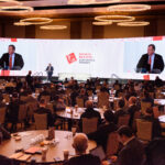 Conference attendees sit at round tables in a large, well-lit room. A speaker stands at a podium on stage with a large screen displaying his image. The screen has a logo for the Sports Betting Executive Summit.