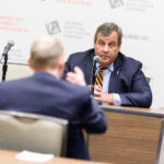 A man in a suit speaks into a microphone at a table, facing another person who is gesturing. The backdrop features logos for the American Gaming Association and the Sports Betting Executive Summit.