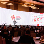 Conference room with attendees seated at round tables, facing a large, curved screen showing the "Sports Betting Executive Summit" logo and charts. A person is speaking at a podium on stage. The room is well-lit with modern chandeliers above.