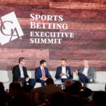 Four men are seated on stage, engaged in a panel discussion at the Sports Betting Executive Summit. The backdrop is a wooden wall with the event logo.