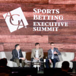 Three men are seated on a stage at the Sports Betting Executive Summit. They are engaged in discussion, with one man speaking into a microphone. The background features a wooden texture with the summit's logo and name.