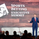 A man in a blue suit and red tie speaks on stage at the Sports Betting Executive Summit. He stands in front of a wooden backdrop with the summit's logo. Chairs and attendees are visible in the foreground.