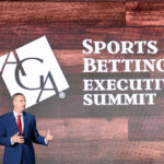 A man in a suit speaks on stage at the Sports Betting Executive Summit. The background features a wooden wall with the summit's name and the AGA logo displayed prominently.