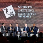 Five men in suits are seated on a stage for a panel discussion at the Sports Betting Executive Summit. A large screen behind them displays the event's name. Audience members are partially visible in the foreground.