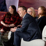 Four people sit on stage in discussion. A man in a suit is in the foreground, with three women seated next to him. They are in a modern setting with a large backdrop and a round table with water bottles.