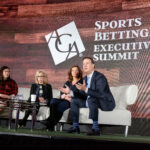 Four people sit on a stage during the Sports Betting Executive Summit. A man in a suit gestures while others, including two women in glasses and one in a red outfit, listen. The background shows a wooden design and the summit's logo.