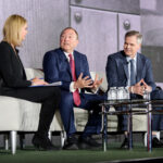 Three people are seated on a stage during a panel discussion. A woman on the left is holding a notepad. The two men in suits are engaged in conversation, with the one in the middle gesturing. They are sitting on a beige couch with a table of water bottles nearby.