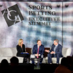Three individuals sit on a stage discussing at the Sports Betting Executive Summit. The backdrop features the conference logo, and the setting includes a light-colored sofa and a small table with water bottles.