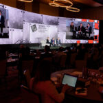Audience members seated at tables watch a panel of speakers on stage at the Sports Betting Executive Summit. The stage features large screens displaying the event's name and live video of the panel discussion.