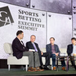 Four men seated on a stage at the Sports Betting Executive Summit, with a grey backdrop displaying the summit's logo. They are engaged in a panel discussion, with two of them holding papers and others listening attentively.