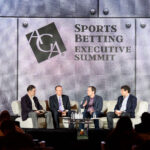 Four men seated on a stage participate in a panel discussion at the Sports Betting Executive Summit. A large sign with the event name is displayed behind them on a metallic backdrop.