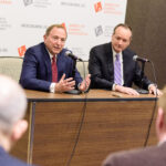 Two men in suits sit at a table with microphones, speaking to an audience at the American Gaming Association's Sports Betting Executive Summit. A logo backdrop and attendees in the foreground are visible.