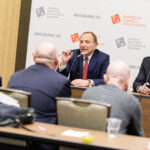 A panel of five men sit at a long table in front of a backdrop with logos for the American Gaming Association and Sports Betting Executive Summit. Two men at the center are speaking, with microphones in front of them.