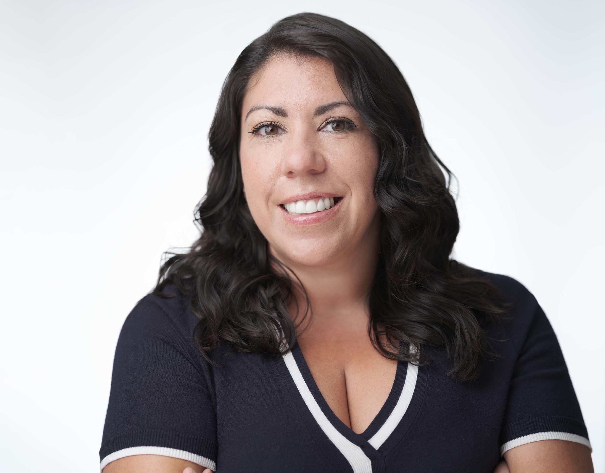 A woman with long dark hair and a friendly smile is wearing a black top with white trim. She has her arms crossed and is posed against a plain white background.