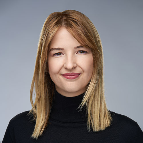 Person with straight, shoulder-length blonde hair, wearing a black turtleneck, smiles gently against a plain gray background.