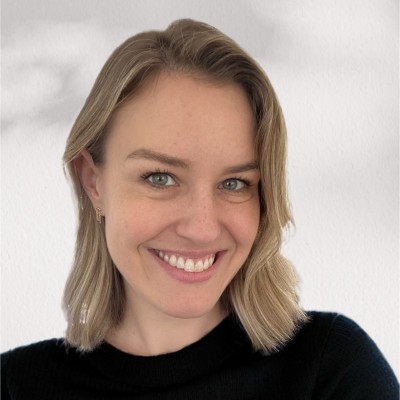 A person with shoulder-length blonde hair and blue eyes, smiling at the camera. They are wearing a black top and standing against a light gray background.