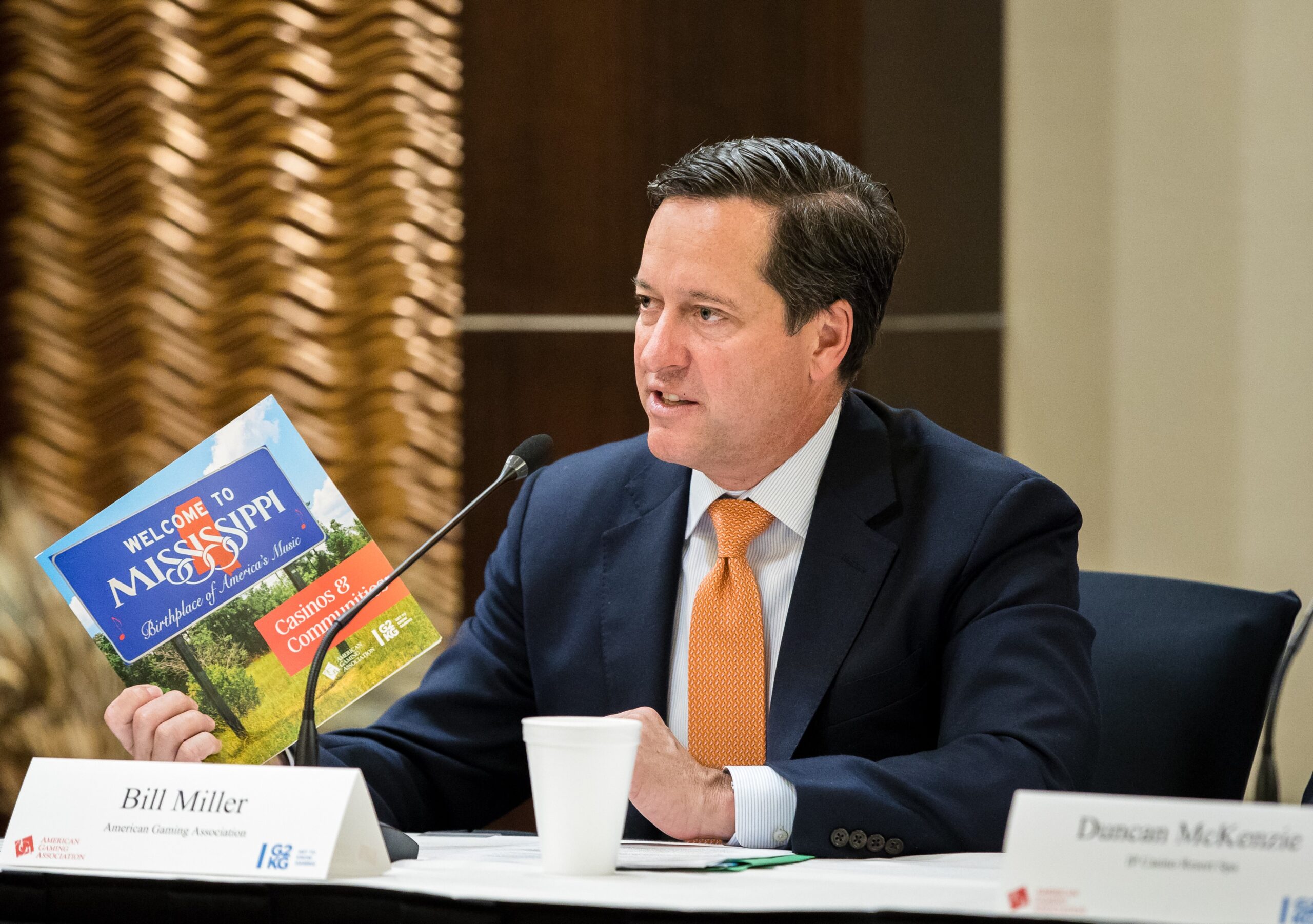 A man in a suit and orange tie speaks at a conference table, holding a "Welcome to Mississippi" booklet. A nameplate reading "Bill Miller" is in front of him. The setting appears formal with a patterned wall and a cup on the table.
