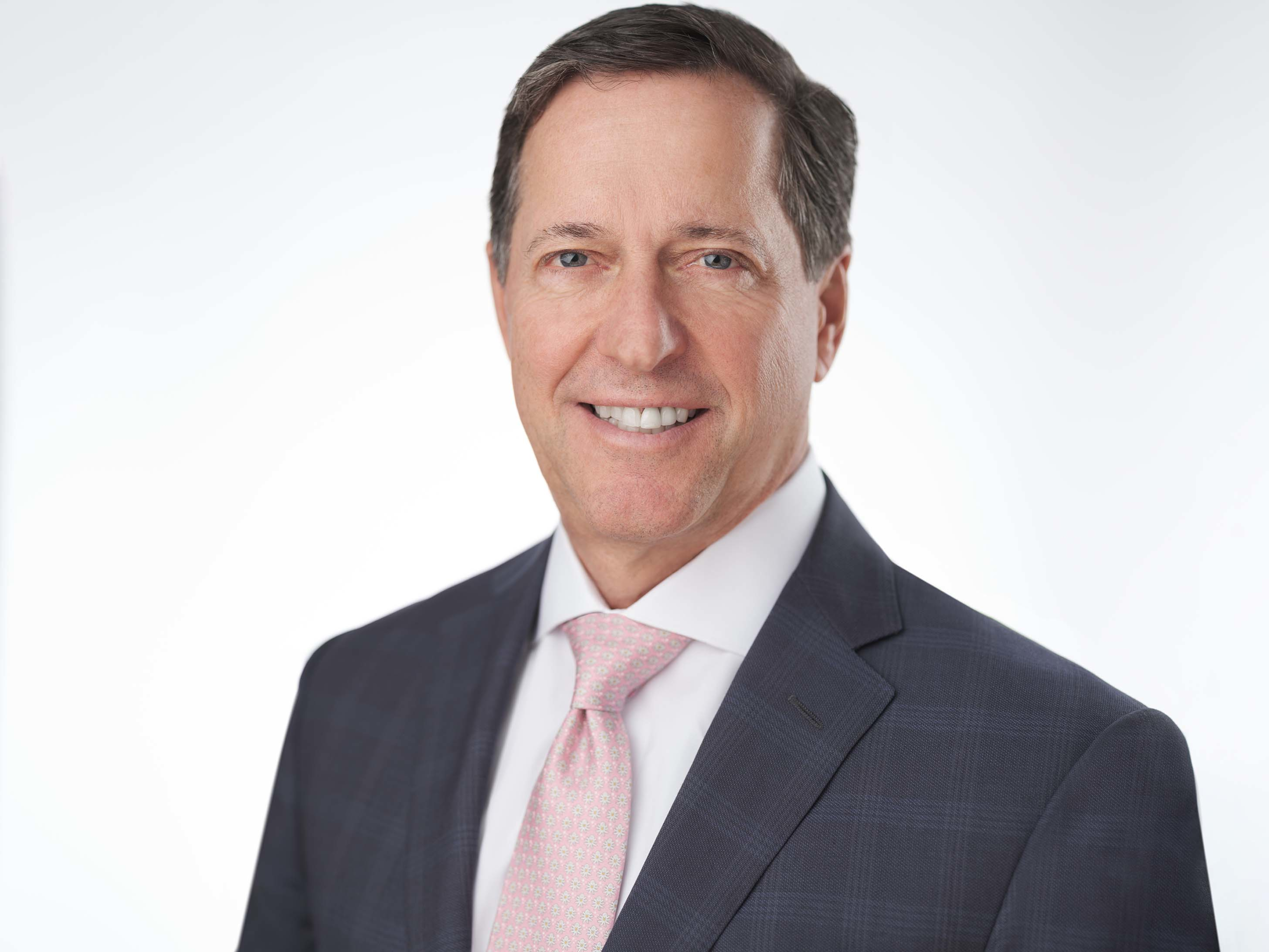A man in a suit with a light pink tie smiles against a plain white background. His hair is dark and he has a light skin tone.