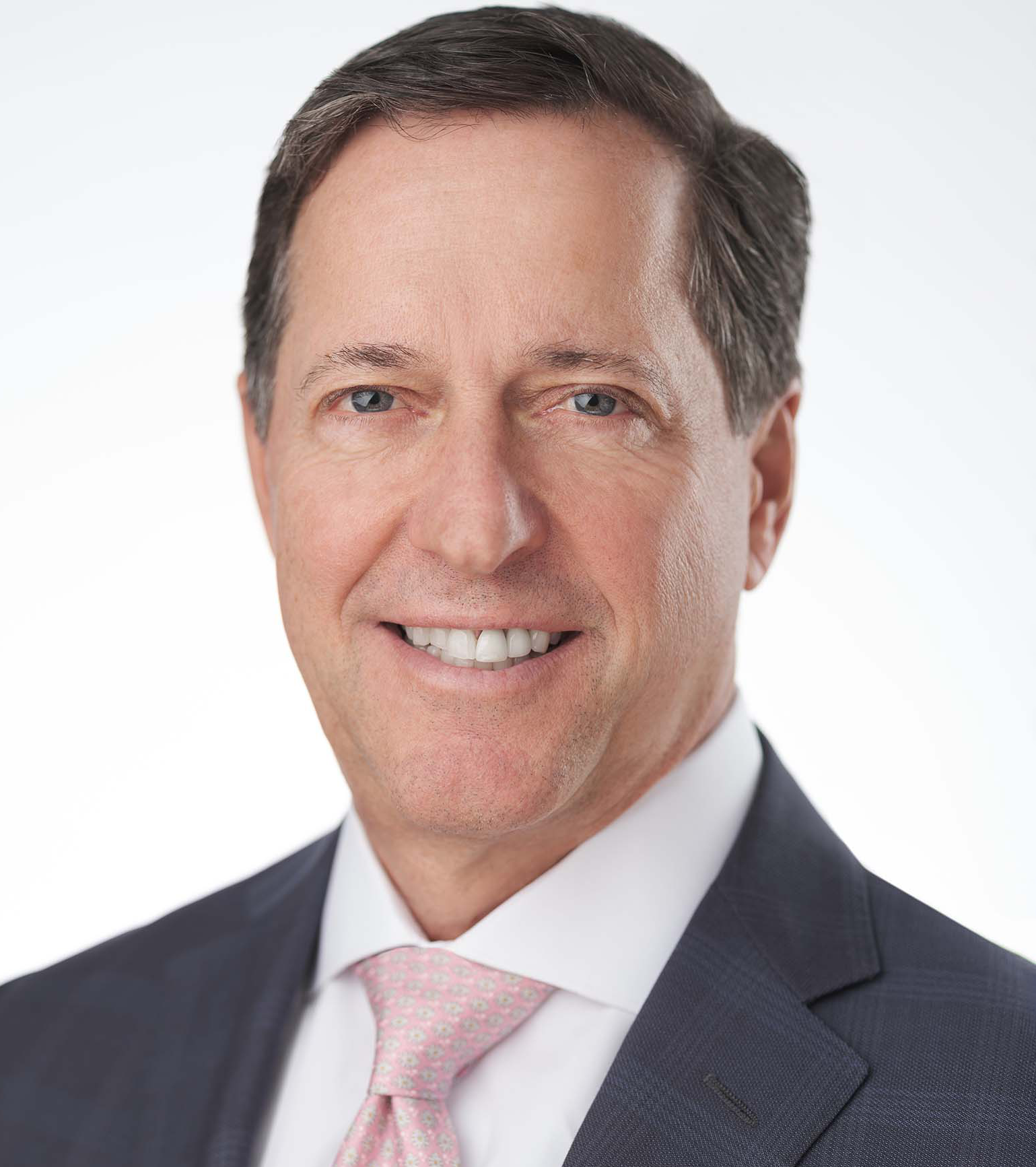 A man in a suit and tie smiles at the camera against a plain background. He wears a gray suit, white shirt, and a pink tie.