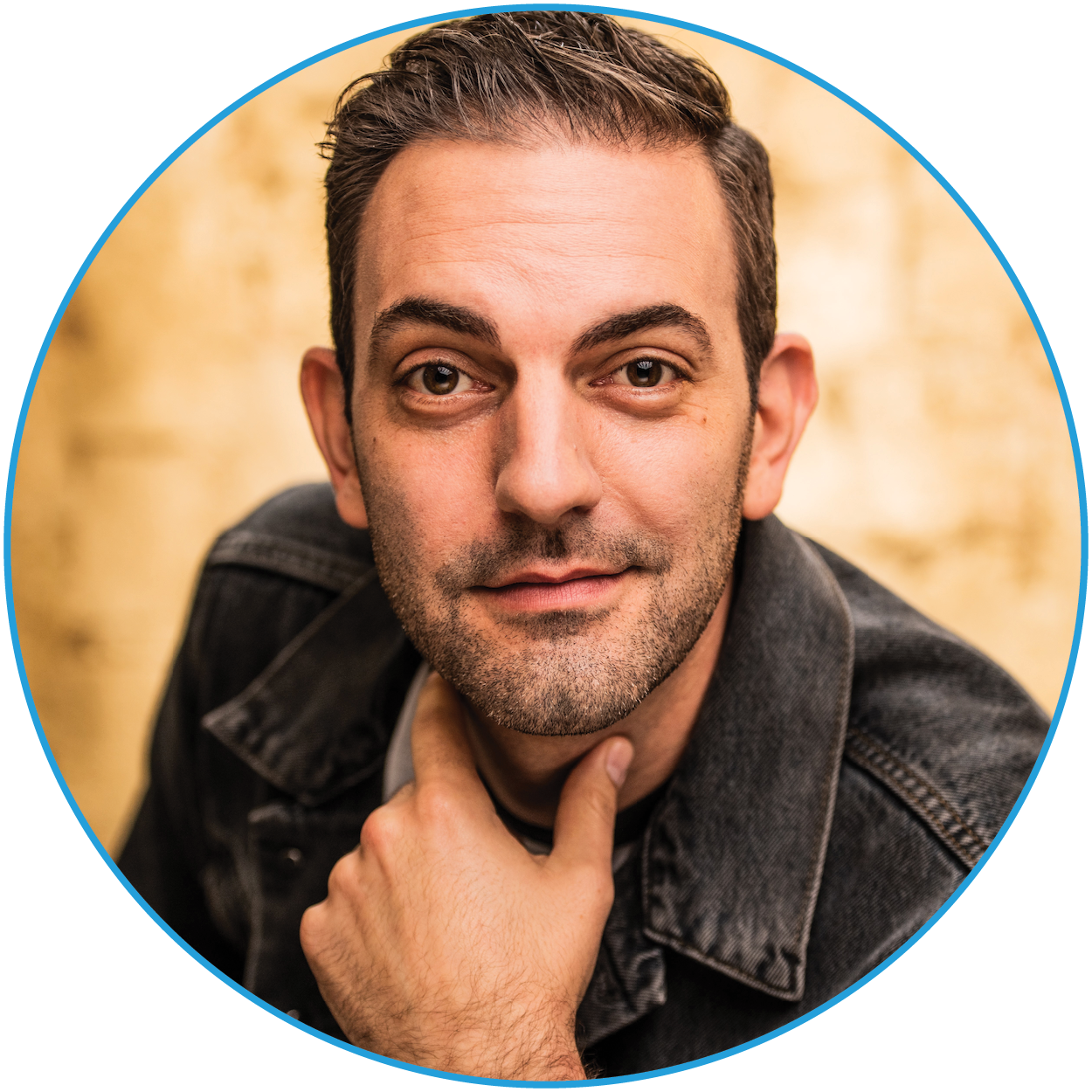Man with short dark hair and a beard, wearing a denim jacket and a white shirt, posing with his hand under his chin. He is looking directly at the camera with a light brown blurred background.