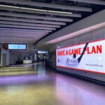 A spacious indoor hallway with a large digital billboard on the right displaying "Have a Game Plan". The left side shows smaller screens. The ceiling has exposed beams and lighting, and the floor is shiny. Some people are visible in the background.