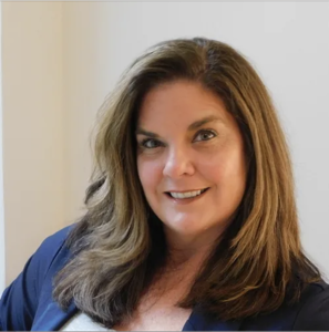 A woman with long brown hair and a navy blazer smiles at the camera against a plain background.
