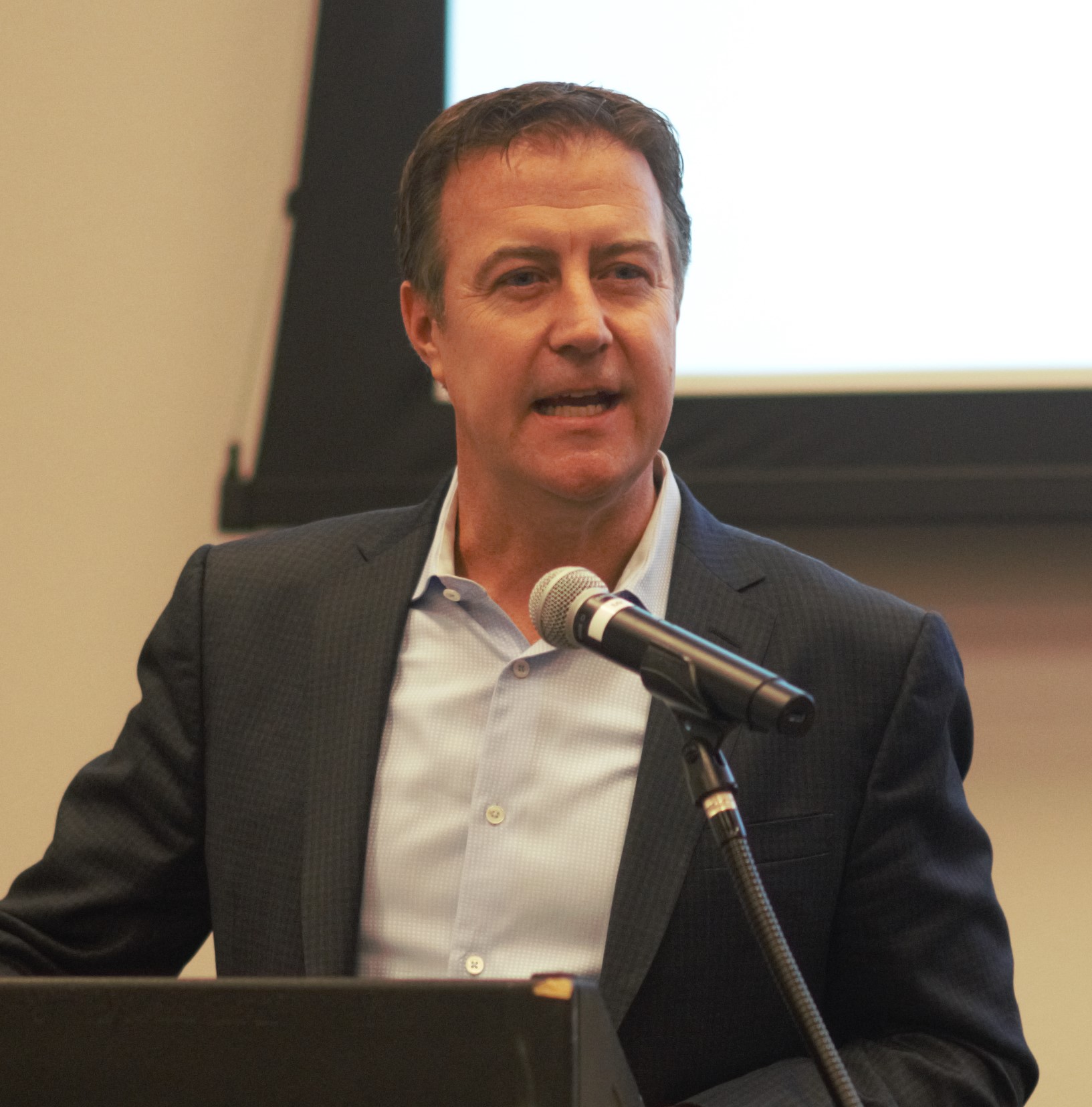 A man in a dark suit and light blue shirt speaks at a podium with a microphone. He is in front of a large screen with a white display, in an indoor setting. His facial expression is engaged, and he is gesturing slightly with one hand.