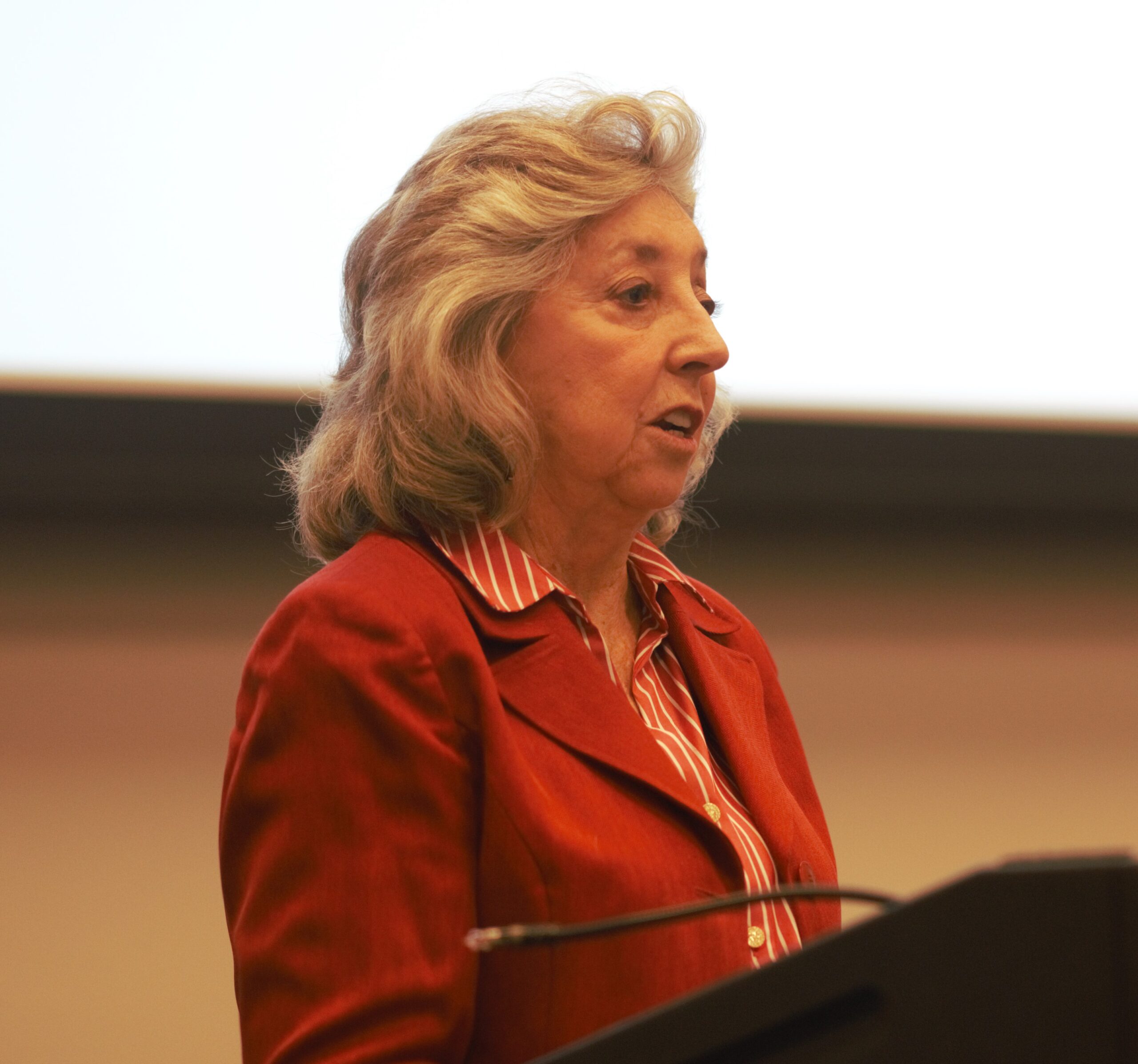 An older woman with wavy gray hair, wearing a red jacket over a striped shirt, is speaking at a podium. The background is mostly white with a dark edge, likely a screen or board.