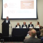 A panel of five people, four seated and one standing at a podium, discuss responsible gaming. Signs for the American Gaming Association and International Gaming Institute are visible on a screen behind them. An audience is seated in the foreground.
