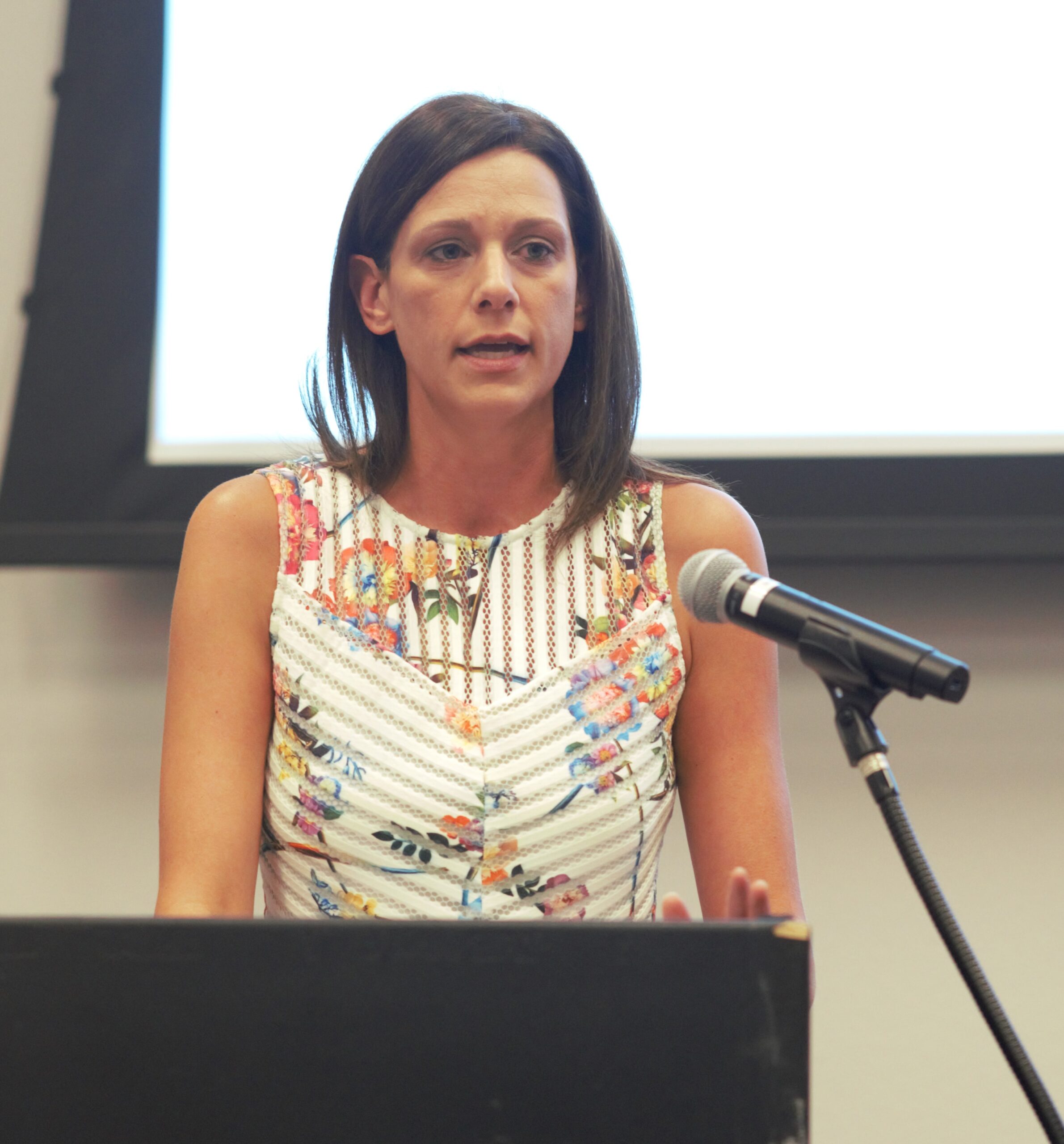 A woman with shoulder-length dark hair stands at a podium, speaking into a microphone. She is wearing a sleeveless, floral-patterned dress. A large, blank screen is visible in the background.