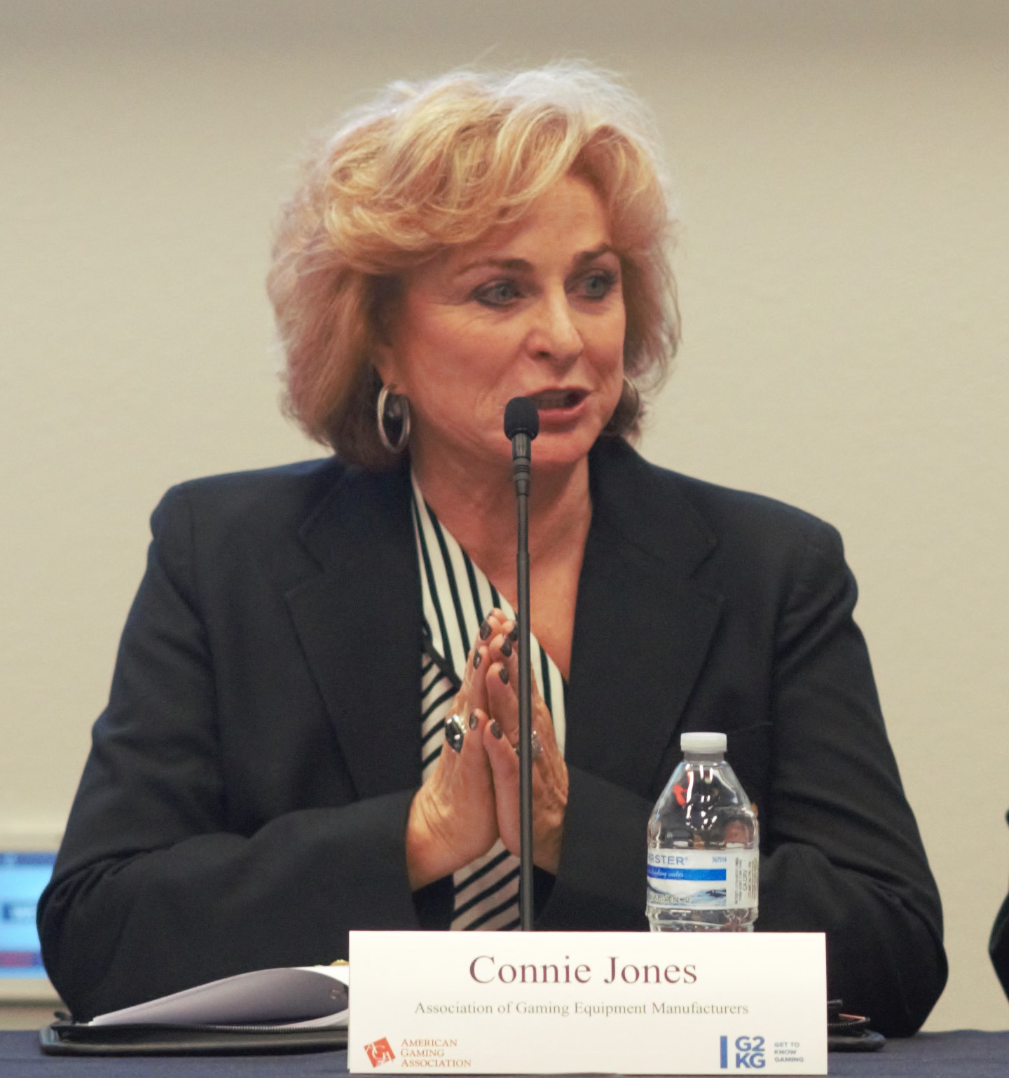 A woman with short blonde hair, wearing a striped blouse and dark blazer, speaks into a microphone at a table. Her hands are clasped. In front of her is a nameplate and a water bottle.