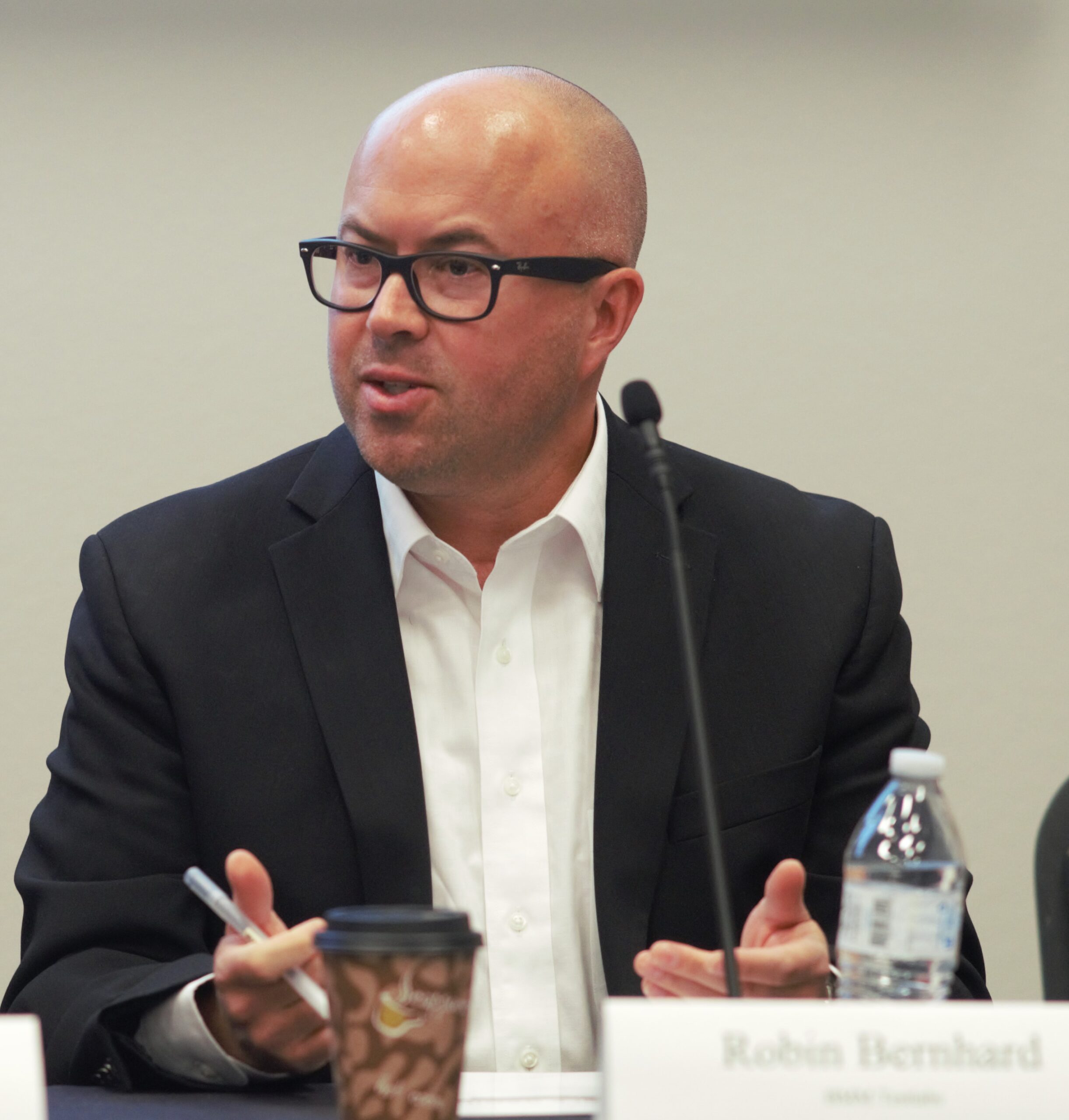 A bald man wearing glasses and a black blazer over a white shirt is speaking while gesturing with his hands. He is seated at a table with a microphone, a disposable coffee cup, and a water bottle in front of him.