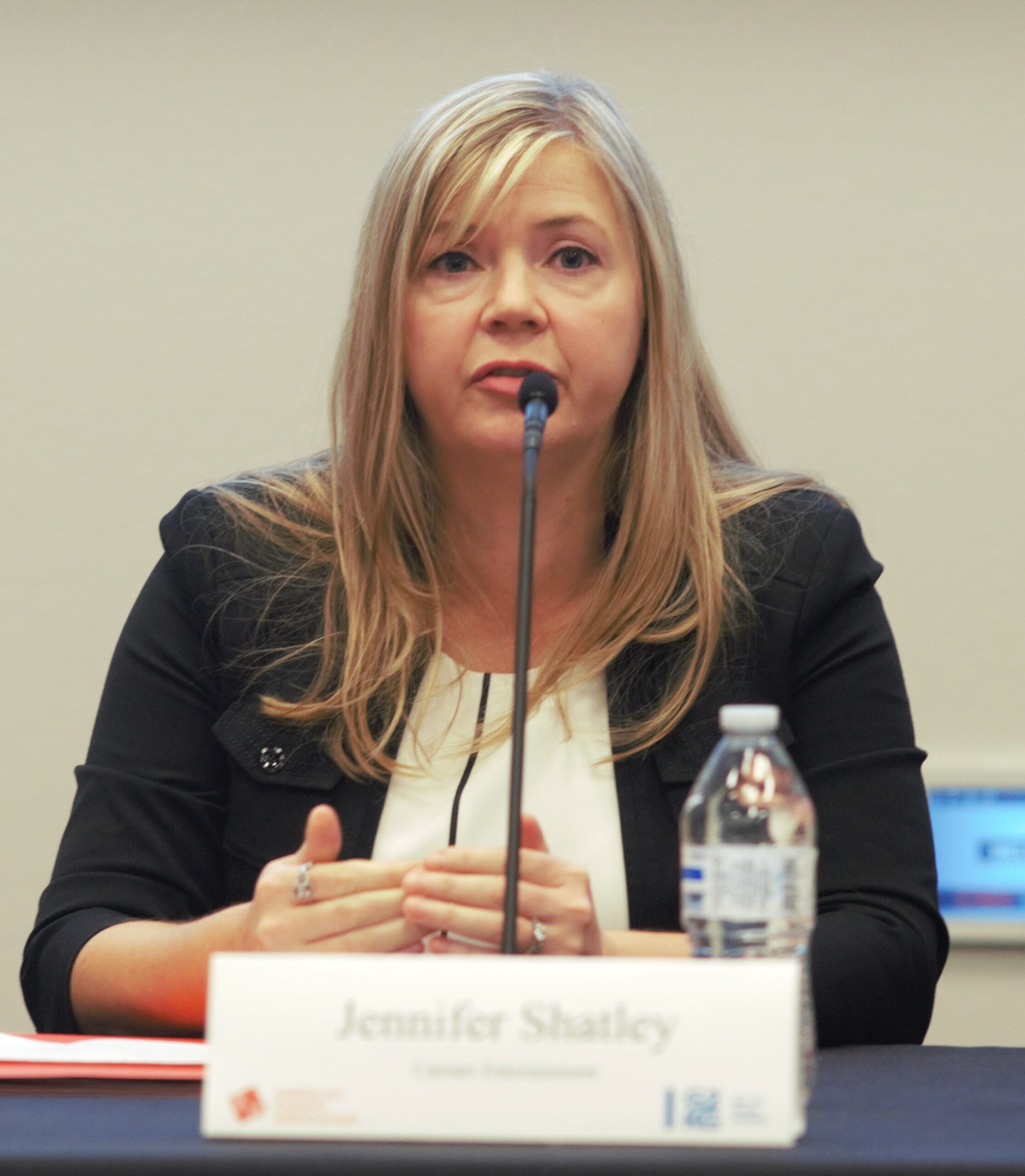 A woman with long blonde hair speaks into a microphone at a table. She wears a black blazer over a white top. A water bottle and a nameplate that reads "Jennifer Shatley" are in front of her.
