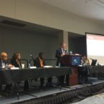 A man speaks at a podium during a conference panel. Four other people sit at a table on the stage, with microphones and papers in front of them. A presentation slide is partially visible in the background.