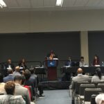 A person stands at a podium speaking to an audience in a conference room. Five panelists are seated at a table on the stage, facing the audience. People are seated in rows, listening attentively.