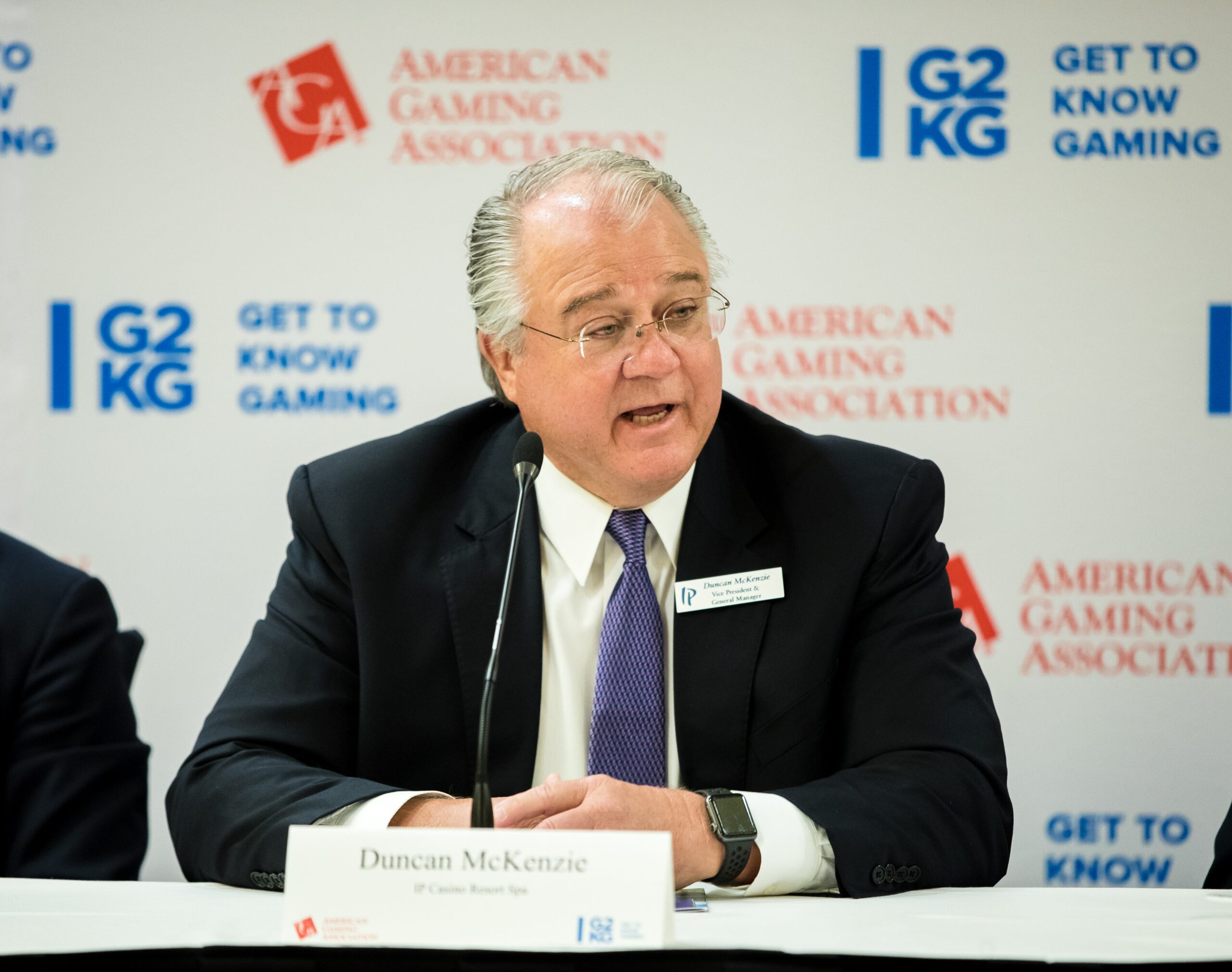 A man in a suit and tie speaks at a panel event, sitting at a table with a microphone in front of him. The background features logos for "American Gaming Association" and "Get to Know Gaming".