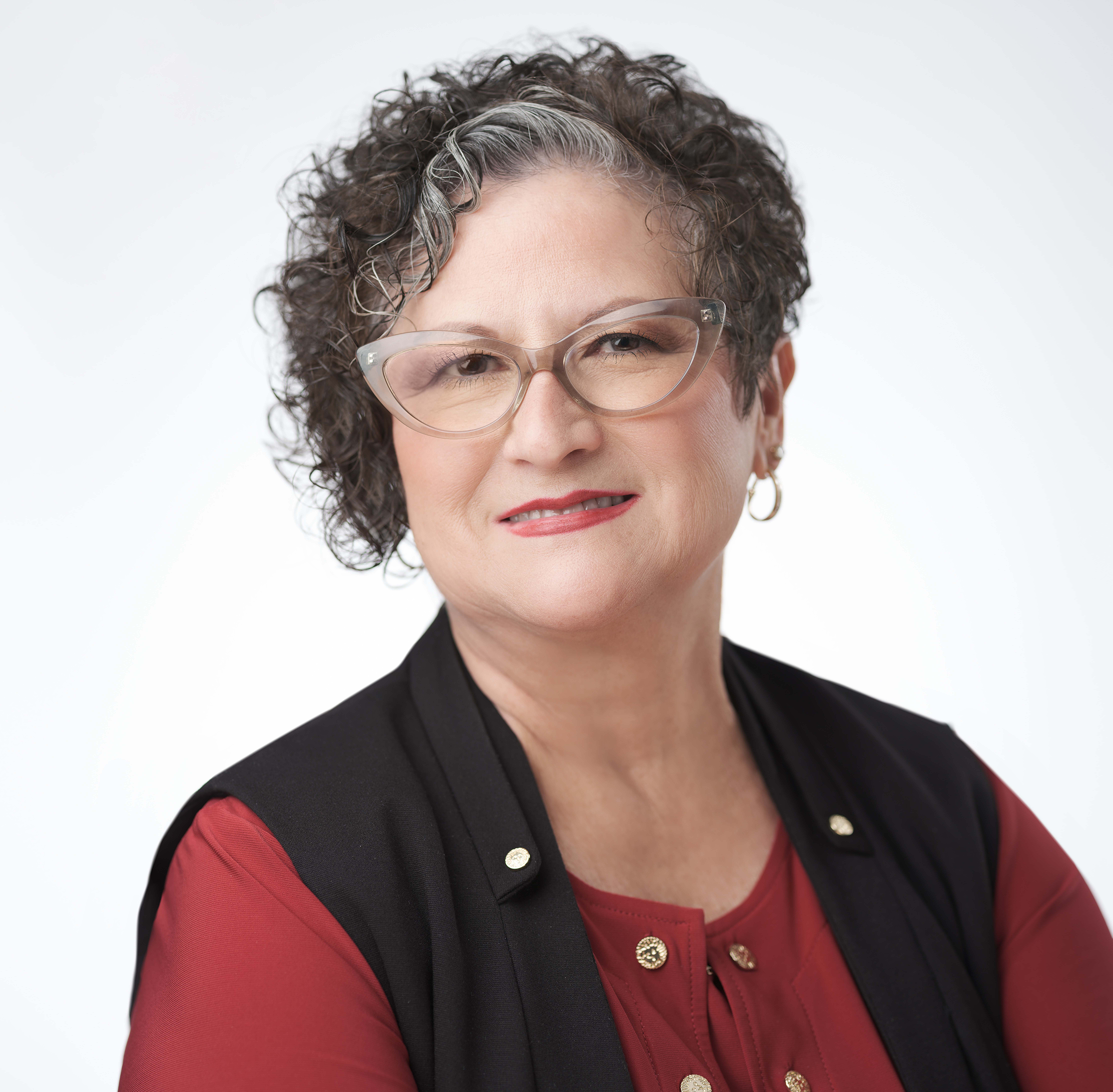 A woman with short curly gray hair and glasses is wearing a red blouse and a black vest. She is smiling slightly against a plain white background.