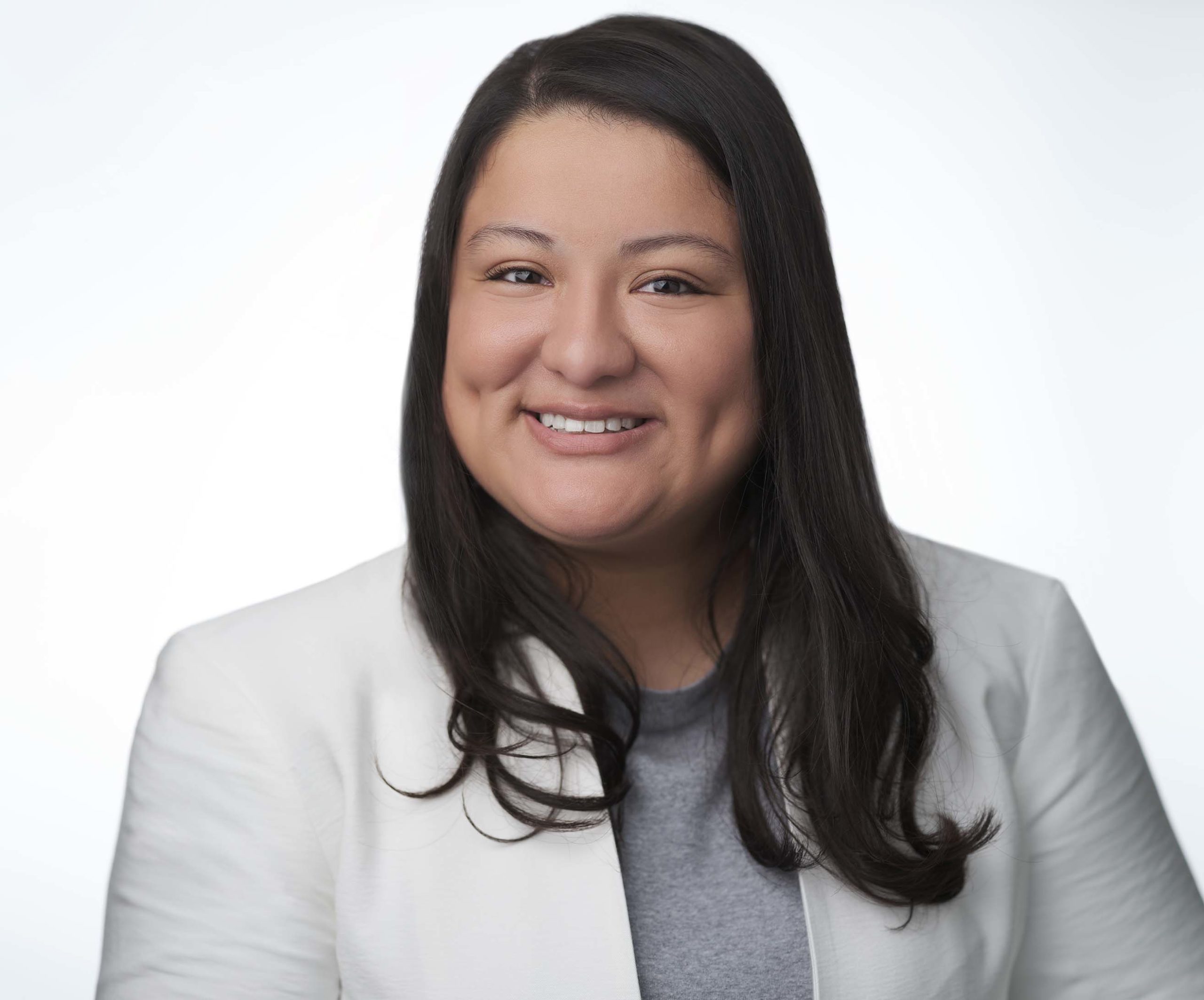 A person with long dark hair smiles, wearing a white blazer over a gray top, against a plain light background.