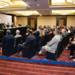 A man addresses an audience at a conference. The room is filled with people seated in rows, listening attentively. A panel of speakers sits at a table on stage. The setting has a modern interior with red carpeting and decorative lights.