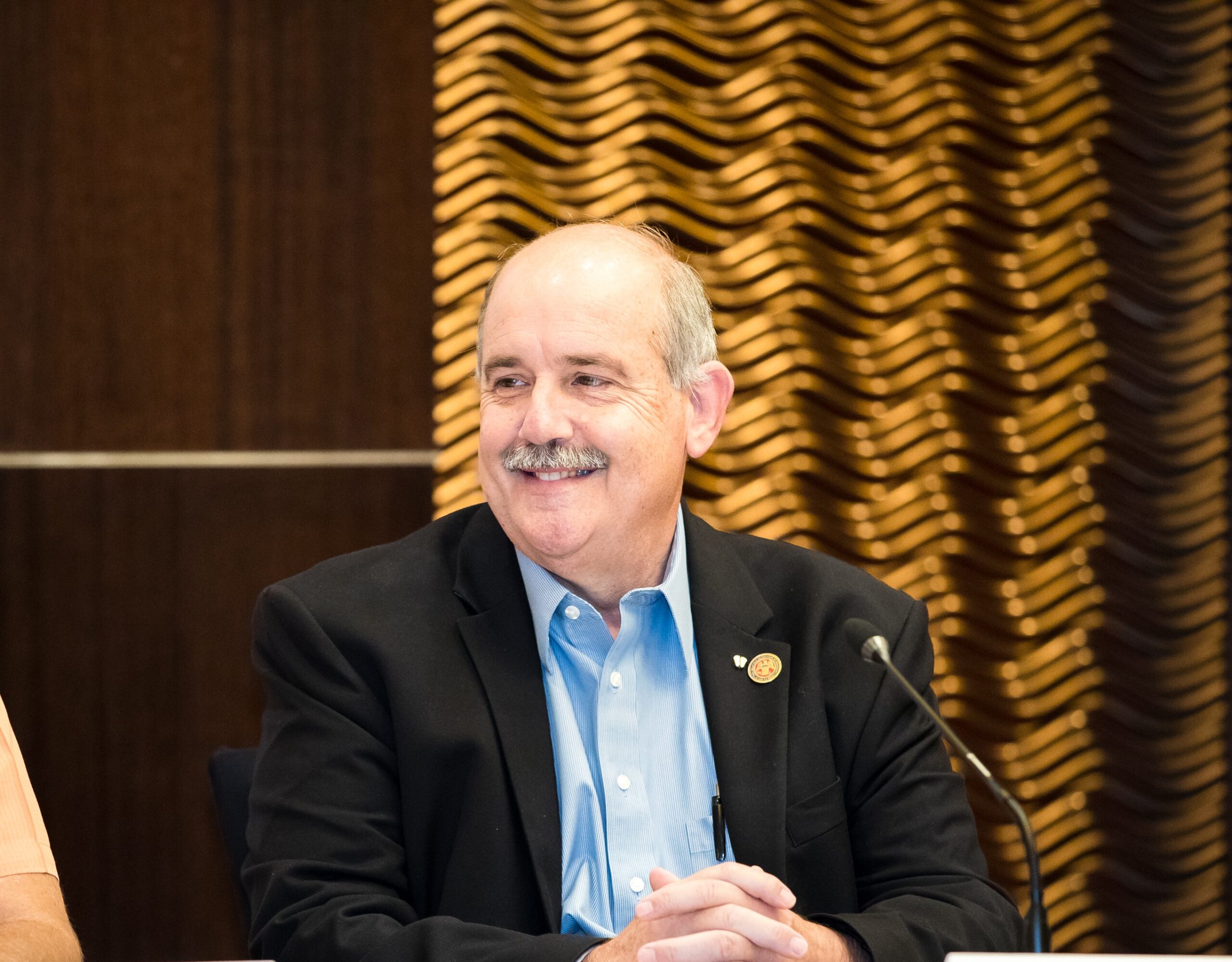 A man with a mustache, wearing a blue shirt and black blazer, is sitting at a table. He is smiling and has a microphone in front of him. The background has a wavy golden pattern on a dark surface.