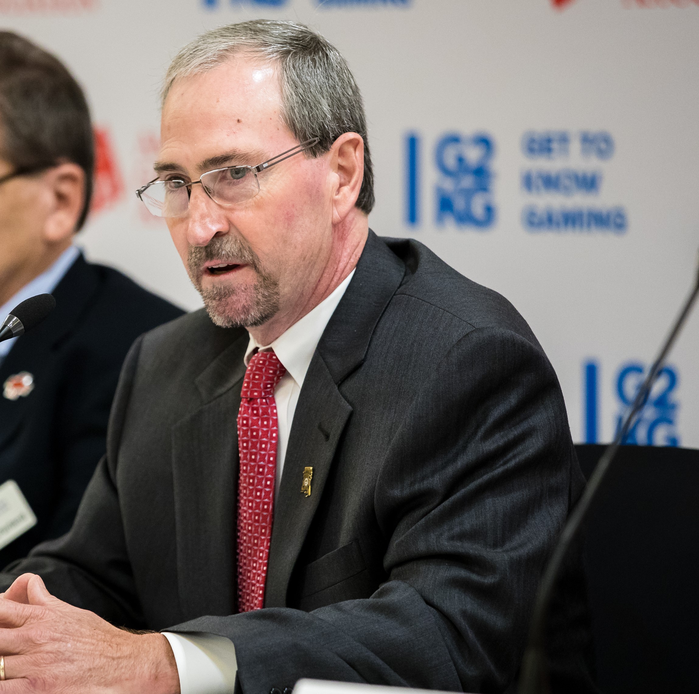 A man in a suit and red tie speaks into a microphone at a conference, sitting at a table with a white banner behind him displaying logos and the text "GET TO KNOW GAMING." Another person is partially visible in the background.