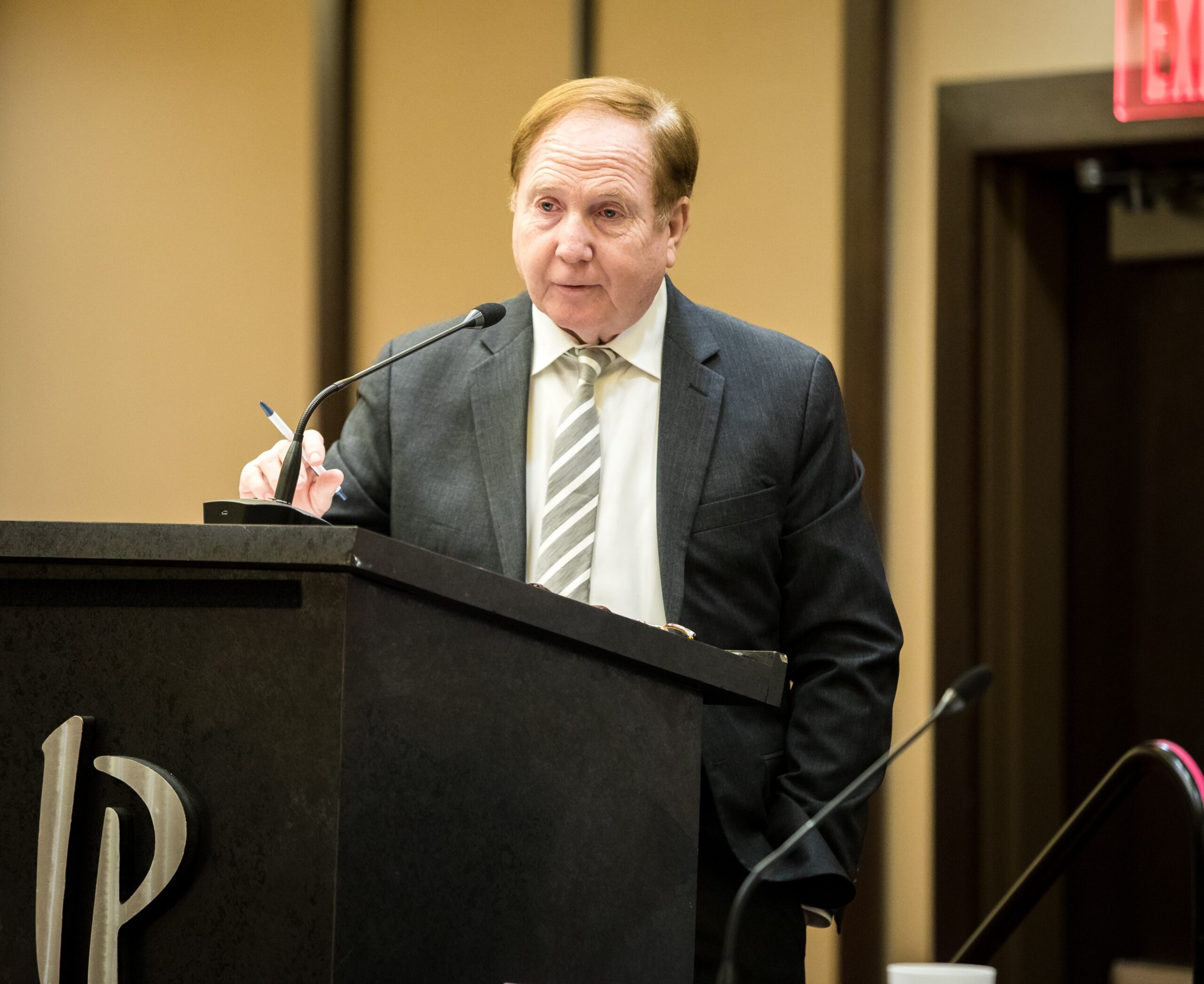 A man in a suit speaks at a podium with a microphone, holding a pen. The podium has a logo, and there is an exit sign visible in the background.