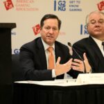 Two men in suits sitting at a table during a press conference. The man on the left is speaking, gesturing with his hands. The backdrop displays logos for the American Gaming Association and "Get to Know Gaming.