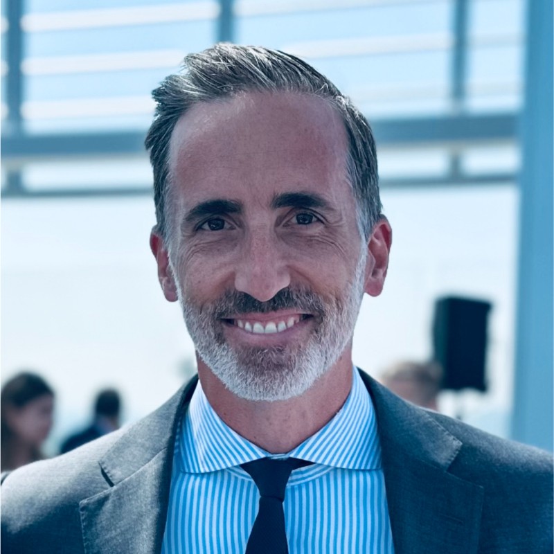 Man in a suit with a blue and white striped shirt and dark tie, smiling outdoors. Background is blurred with architectural details and people.