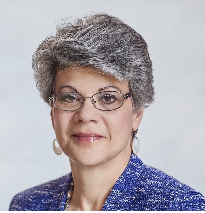 A person with short gray hair and glasses is wearing a blue textured blazer and white earrings. They have a neutral expression and are posed against a light background.