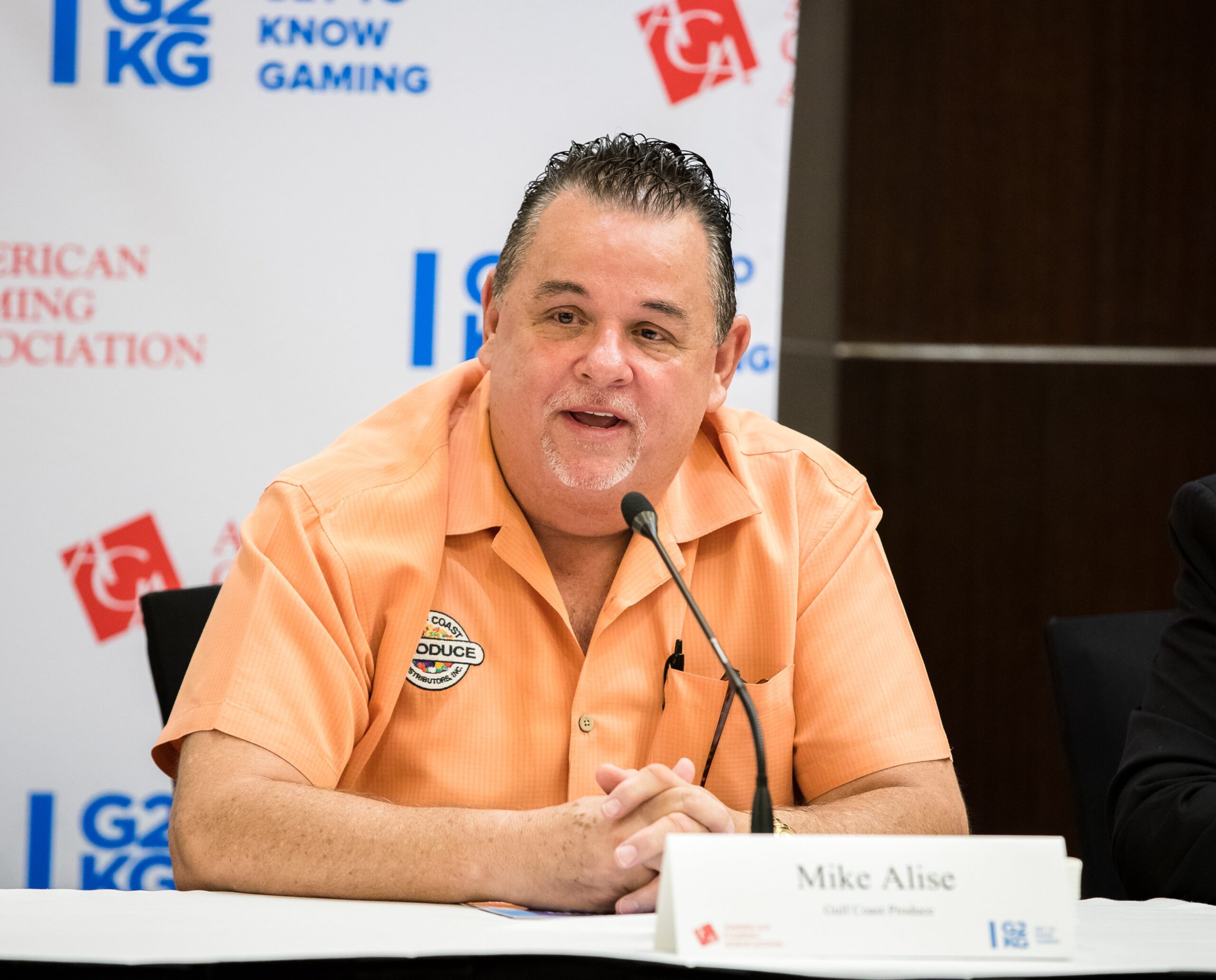 A man in an orange shirt speaks into a microphone at a conference. He's seated at a table with a nameplate labeled "Mike Alise." Various conference logos are displayed on a banner in the background.