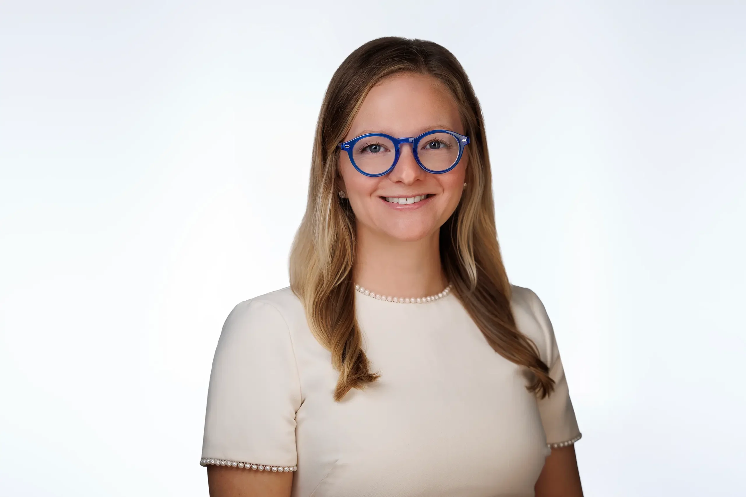 Smiling woman with long blonde hair wearing blue glasses and a cream-colored shirt with pearl-trimmed sleeves, against a plain white background.