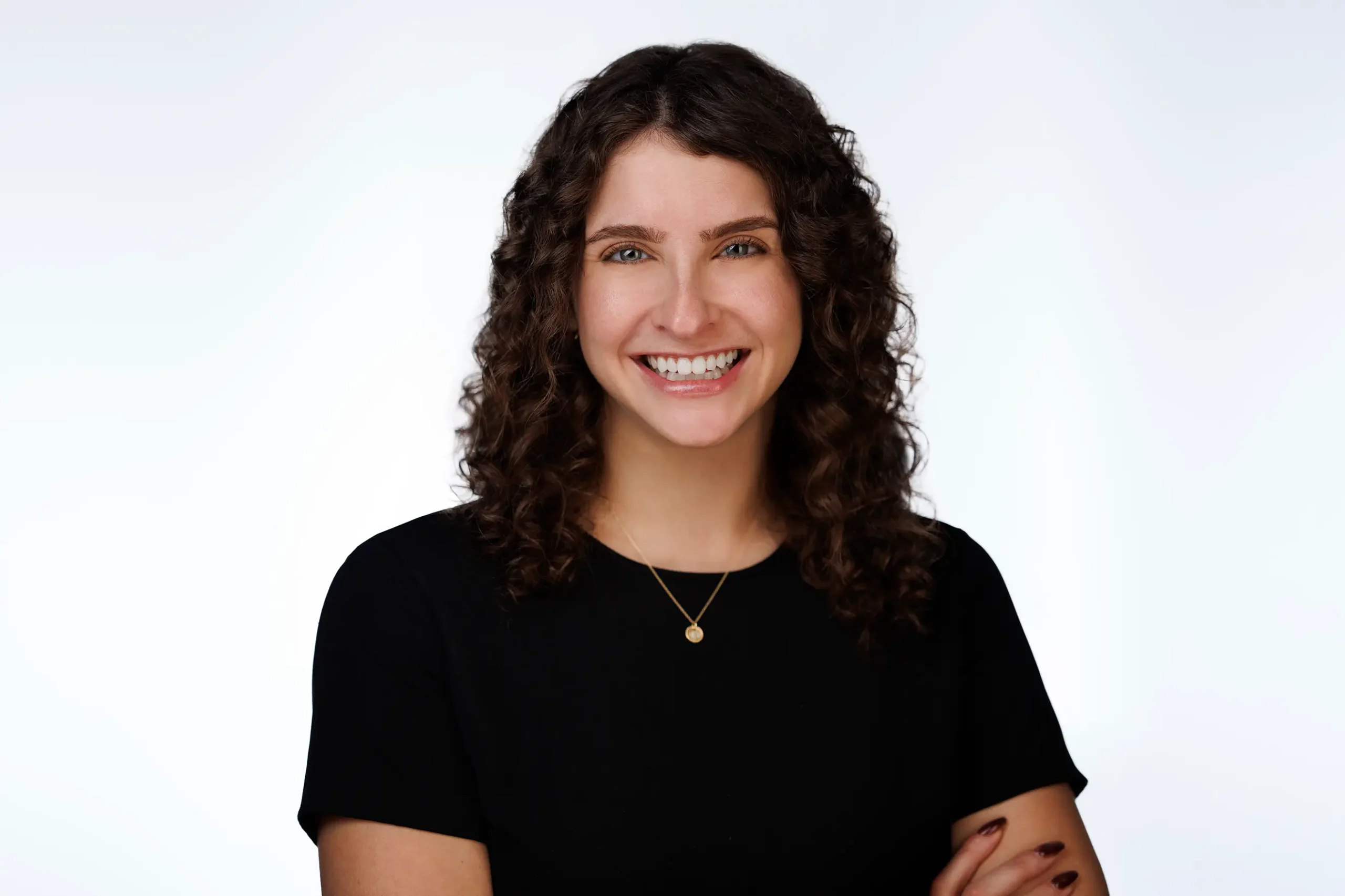 A person with curly brown hair is smiling at the camera. They are wearing a black top and a small necklace, standing against a plain white background. Arms are crossed casually.