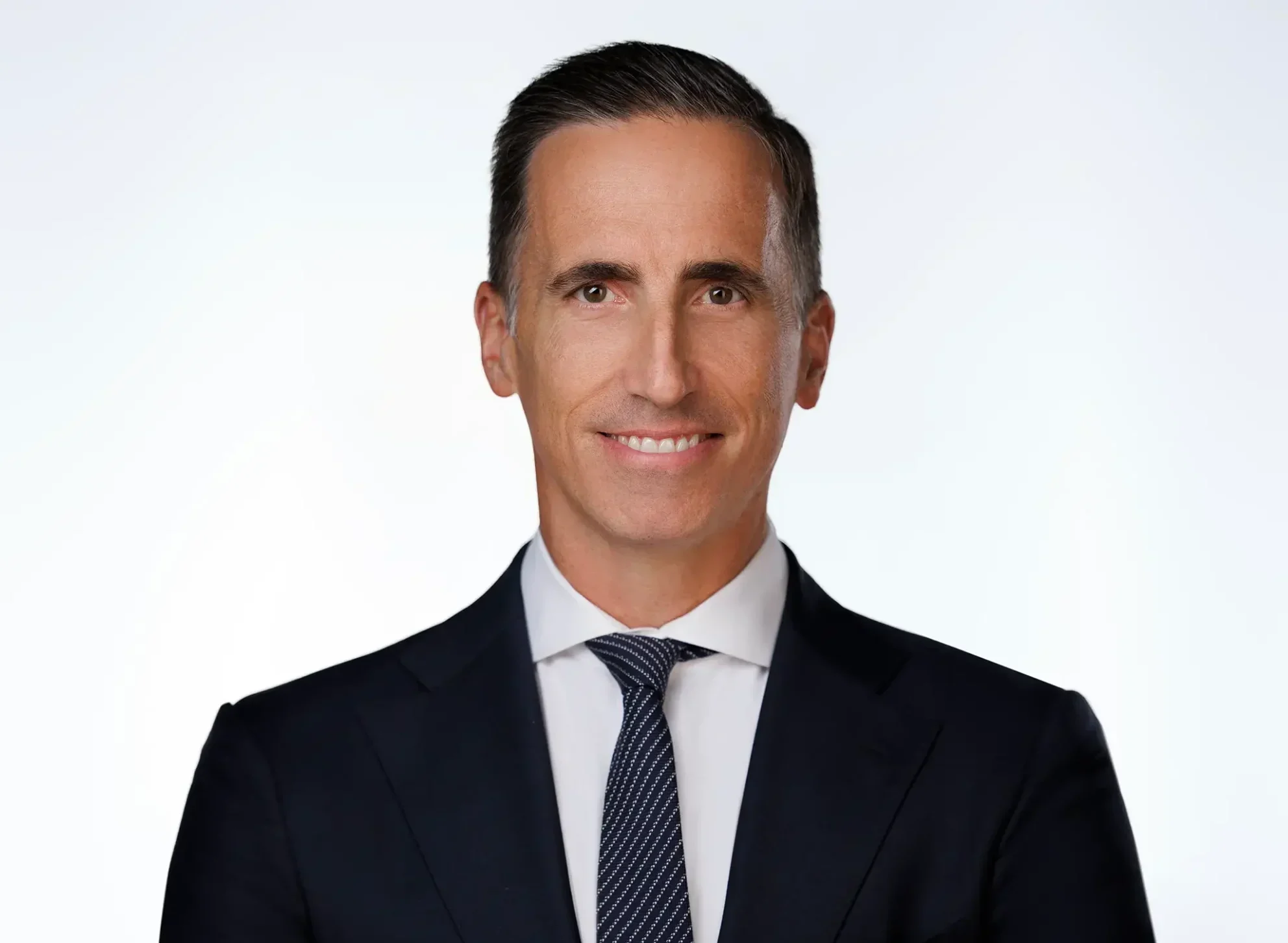Smiling man in a dark suit and tie with short, neatly combed hair, standing against a plain white background.